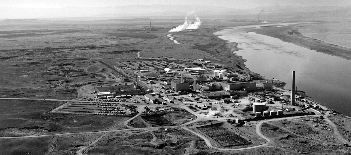 A black and white arial photo of a cluster of buildings and smoke stacks along a river.