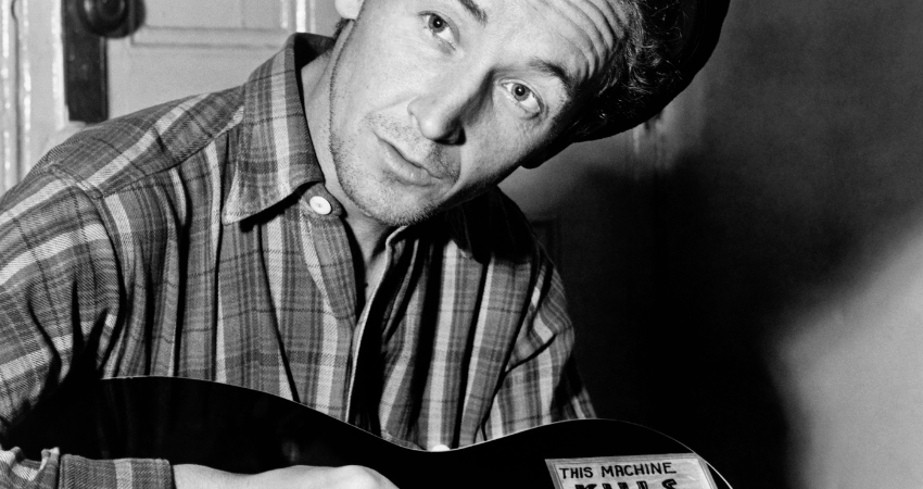 A back and white photograph of Woody Guthrie with his guitar